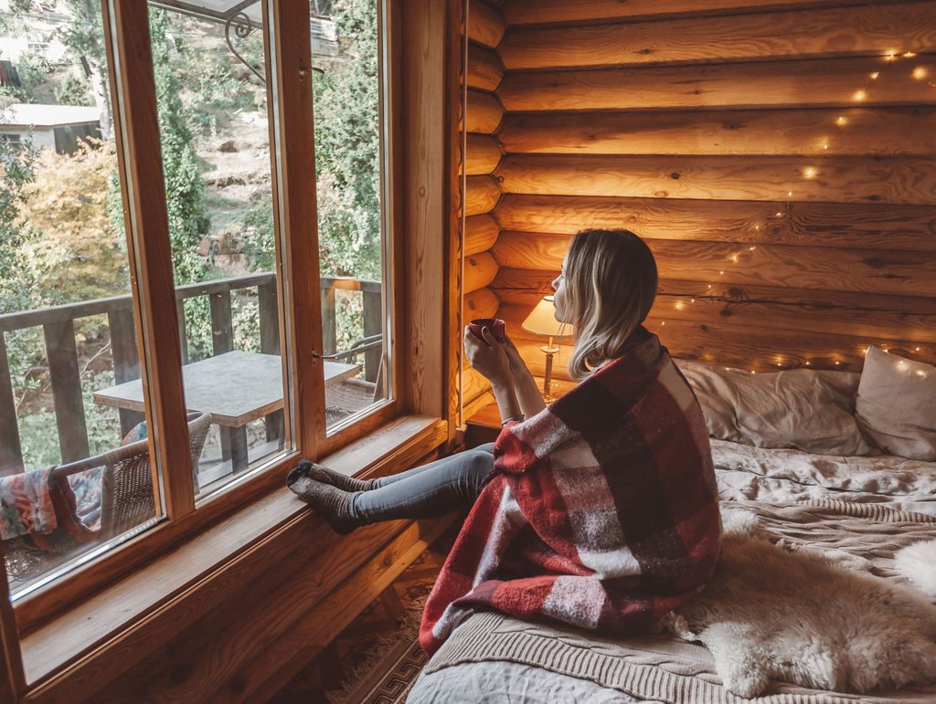 Junge Frau mit Teetasse in warmem Blockhauszimmer sieht aus dem Fenster